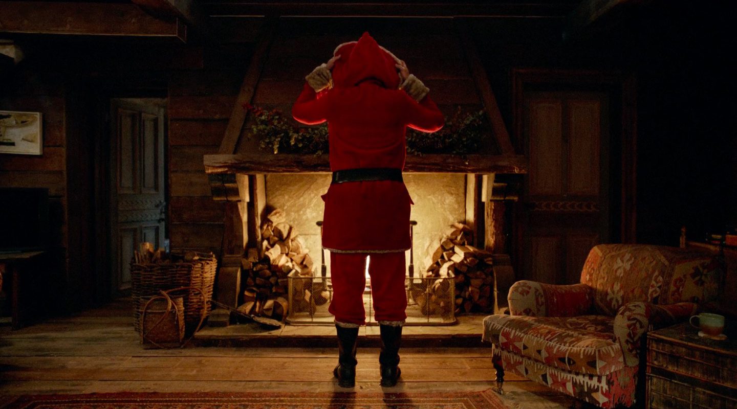 A figure in a red suit at a lit fireplace, seen from behind